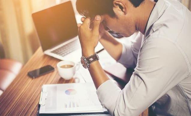 Stressed person sitting at a table with a laptop, coffee, and documents.